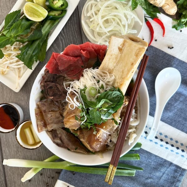 A large bowl of Vietnamese pho with beef, noodles, herbs, and a bone marrow, surrounded by fresh garnishes.
